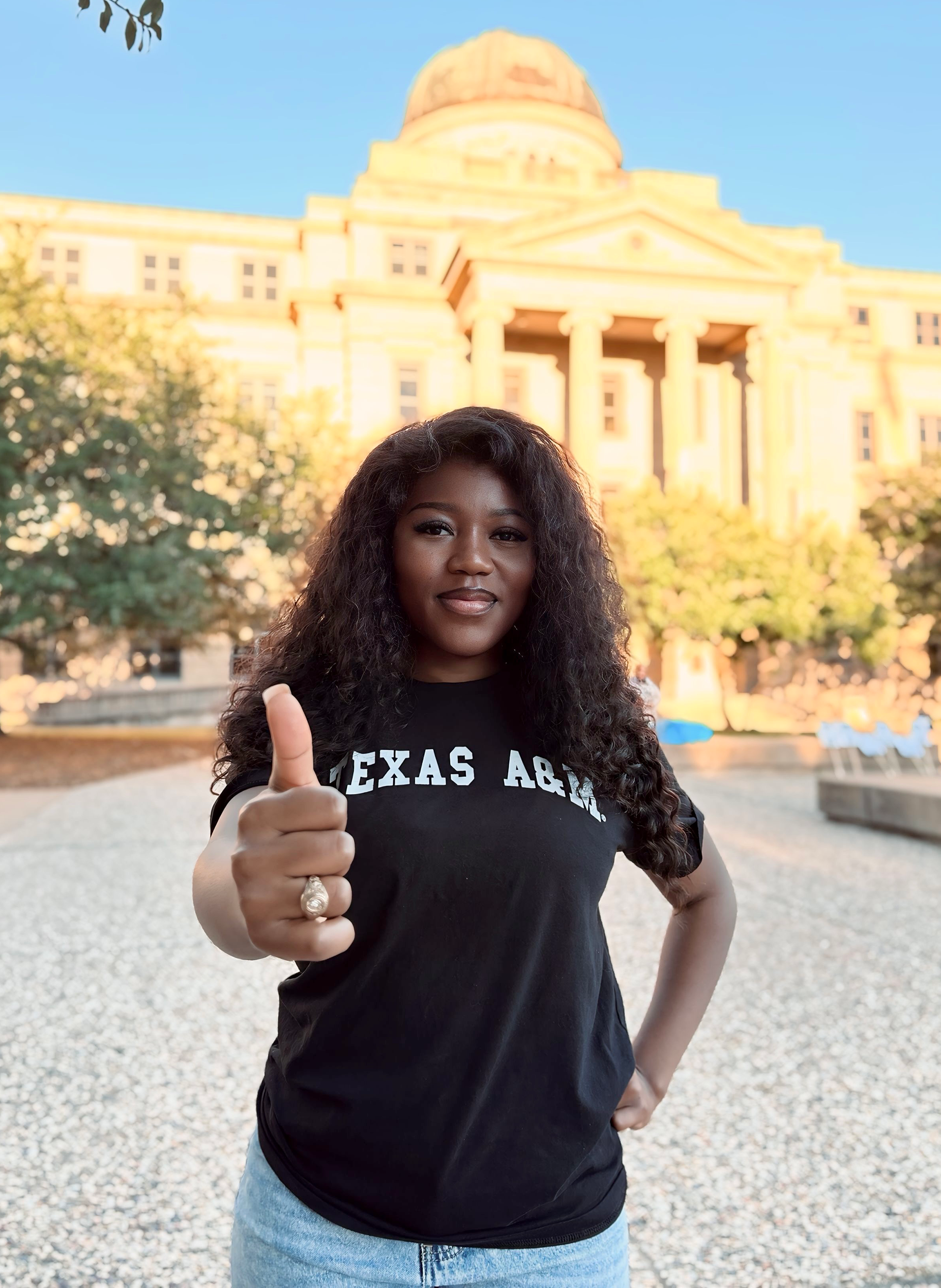 Jewel Fanyui wearing a Texas A&amp;M University T-shirt gives a thumbs up in front of the Academic building.