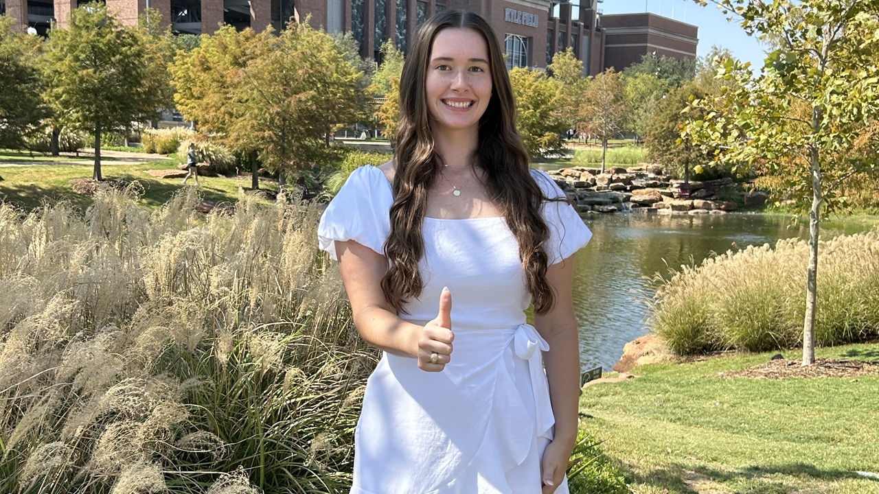 Kailey Rettmann is in a white dress giving a thumbs up, standing in Aggie Park with grass and a pond, with Kyle Field visible in the background.