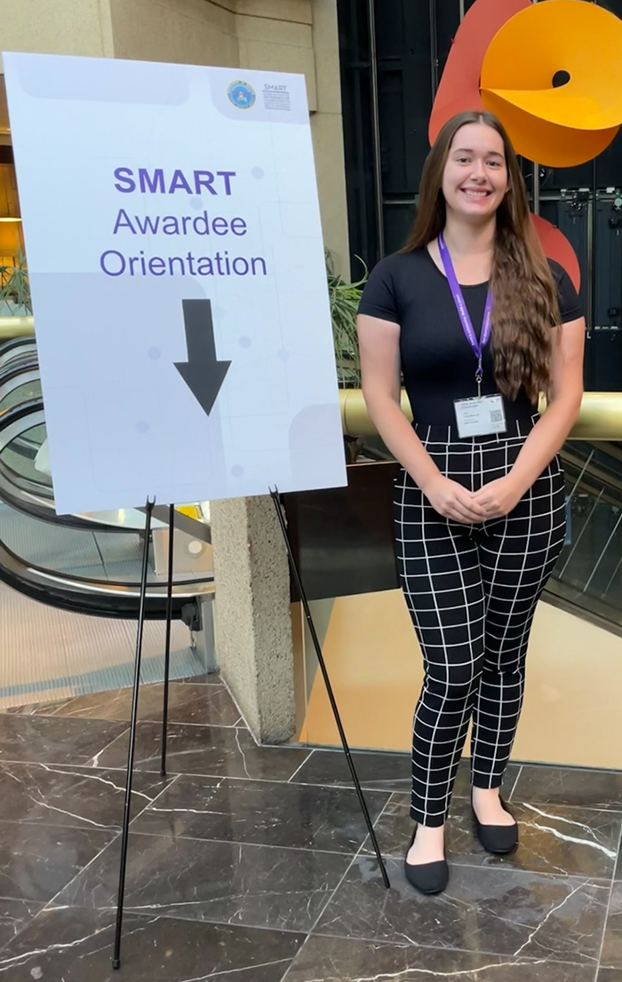 Kailey Rettmann person stands smiling next to a sign that reads "SMART Awardee Orientation," with an arrow The setting appears to be inside a building with artistic decor.