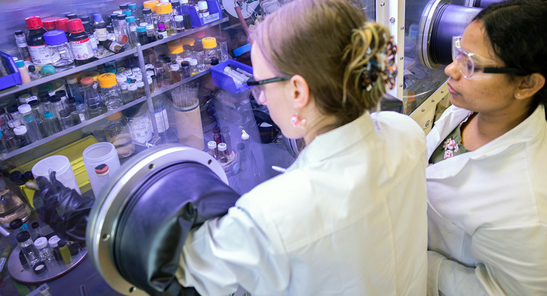 Two scientists working in a laboratory, one viewing a sample in a glove box and the other observing. The lab setting includes various scientific equipment and containers.