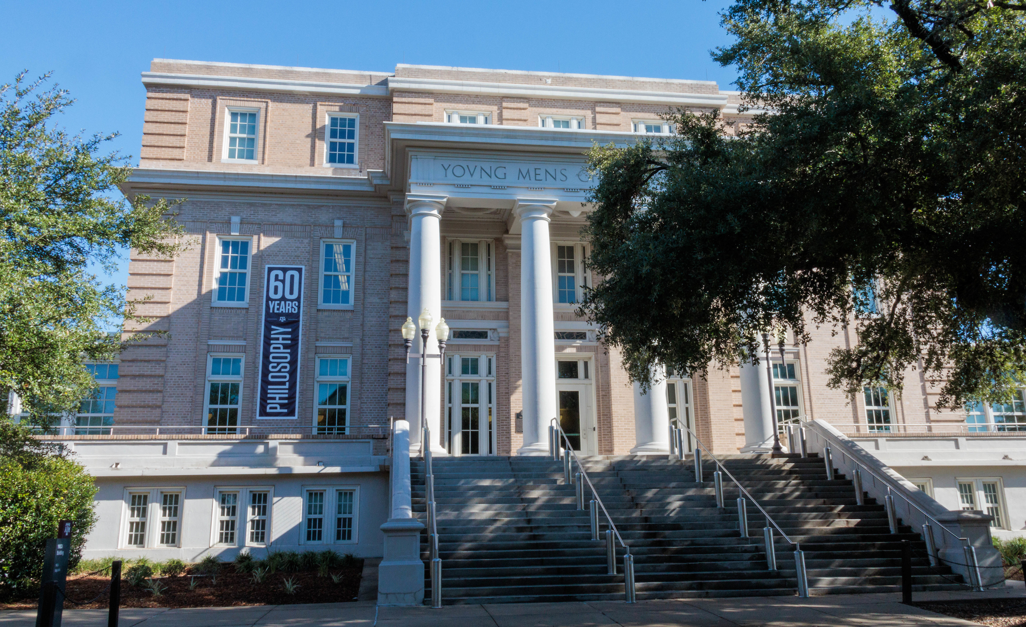 Exterior view of the Young Men’s Christian Association building on the campus of Texas A&amp;M with a banner celebrating 60 years of philosophy on the left side of the building.