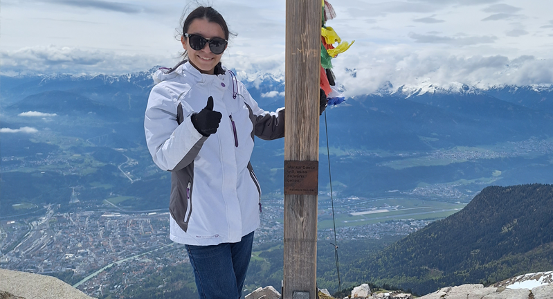 Juliette Rocha standing next to a wooden post with prayer flags, giving a thumbs-up, with a panoramic view of a mountain range and a city below in the background.