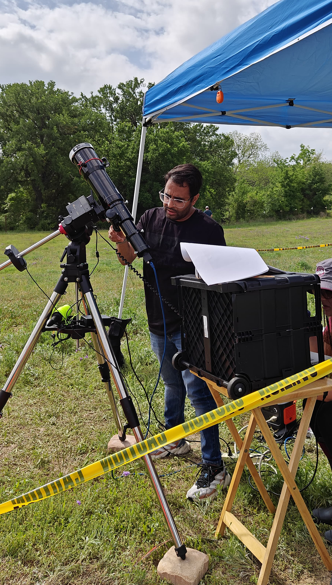 Sahir Gagan is adjusting a telescope mounted on a tripod in a field with a canopy and equipment nearby.