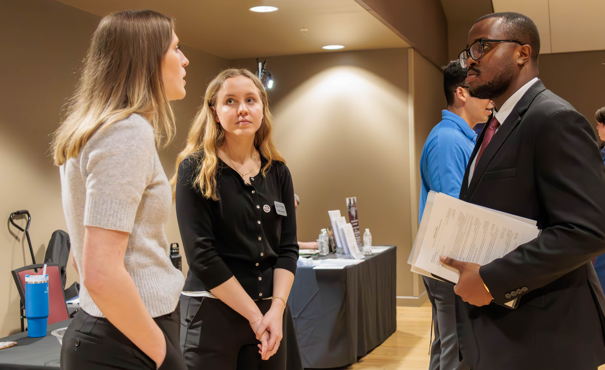 Two representatives from Trenegy speak with an Economics student dressed in a suit and carrying a folder.