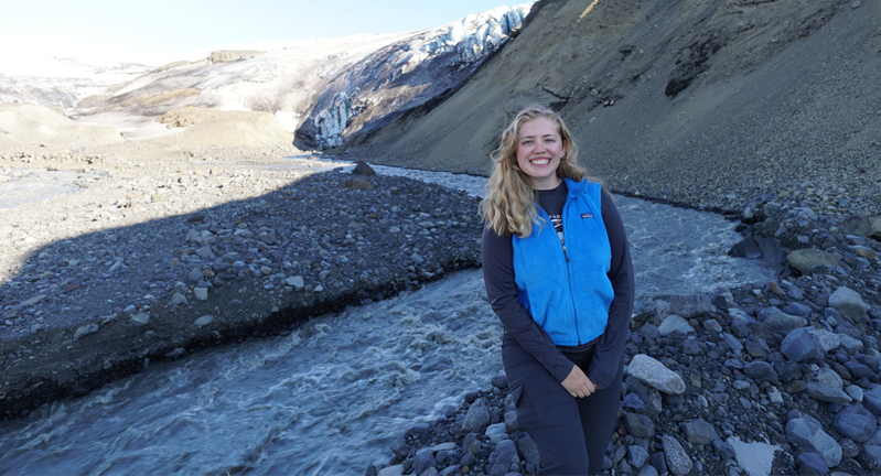 Lauren Berger is standing in a blue jacket smiling in front of a scenic view with a flowing river and a glacier in the background.