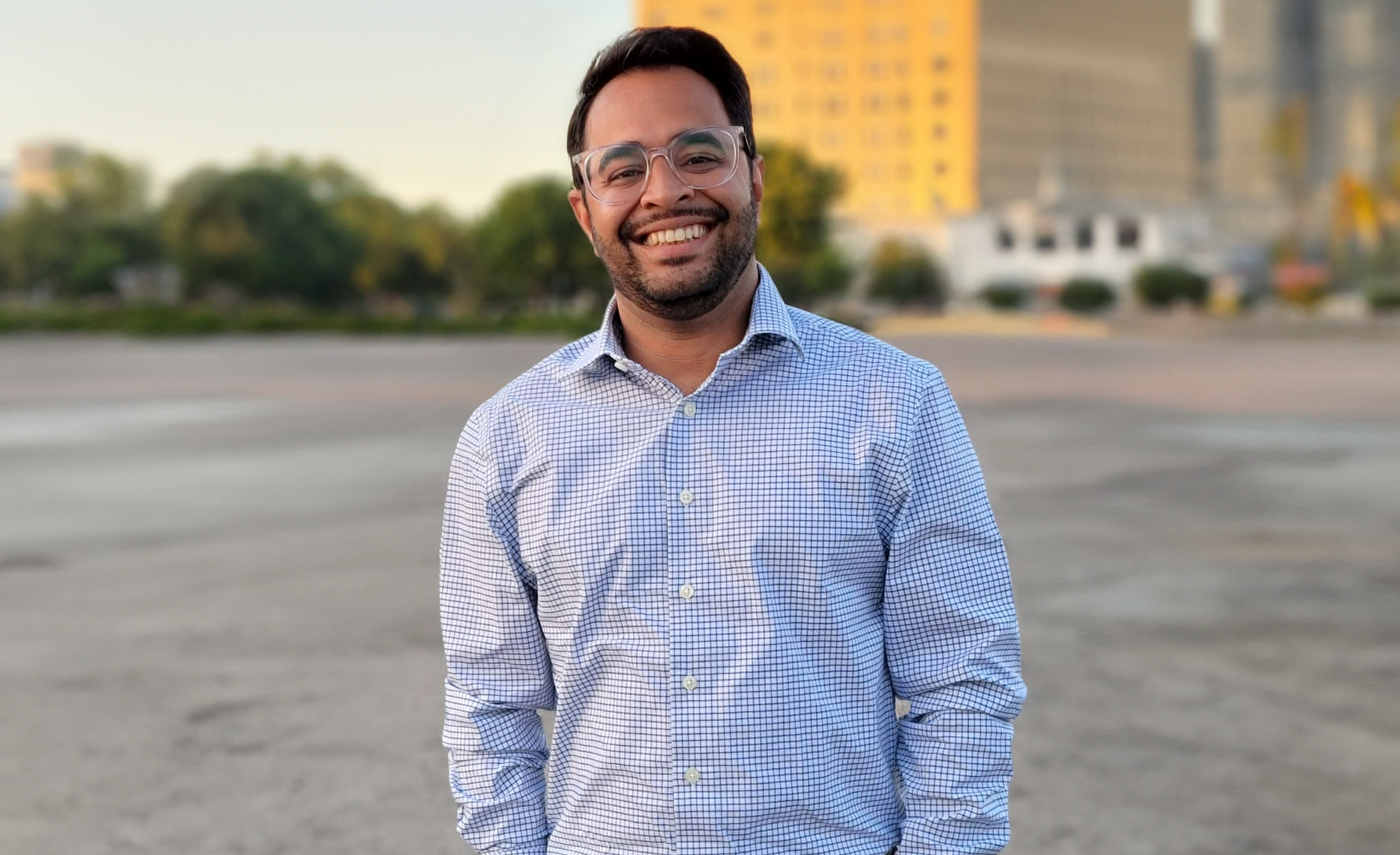 Sahir Gagan smiling with glasses, wearing a blue checkered shirt, standing outdoors with city buildings in the background.