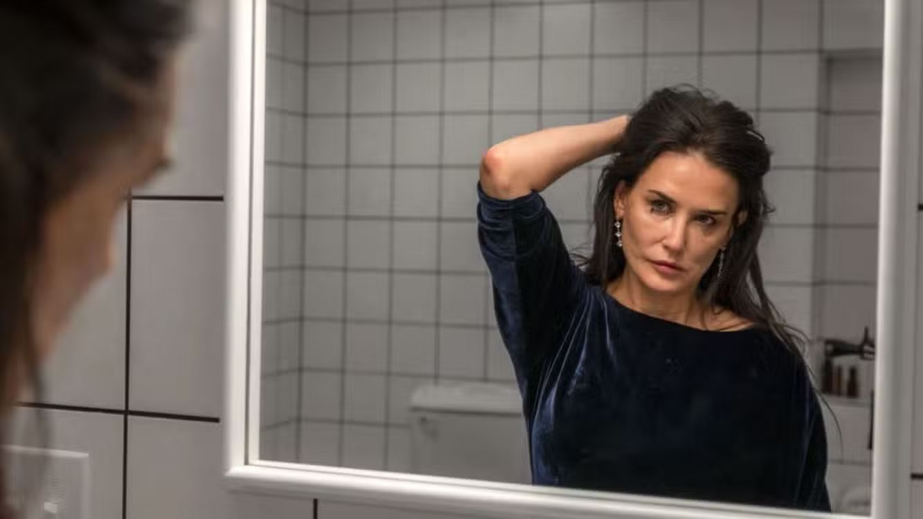 A person is looking at their reflection in a bathroom mirror, fixing their hair. They are wearing a blue top. The background features a simple, white tiled bathroom.