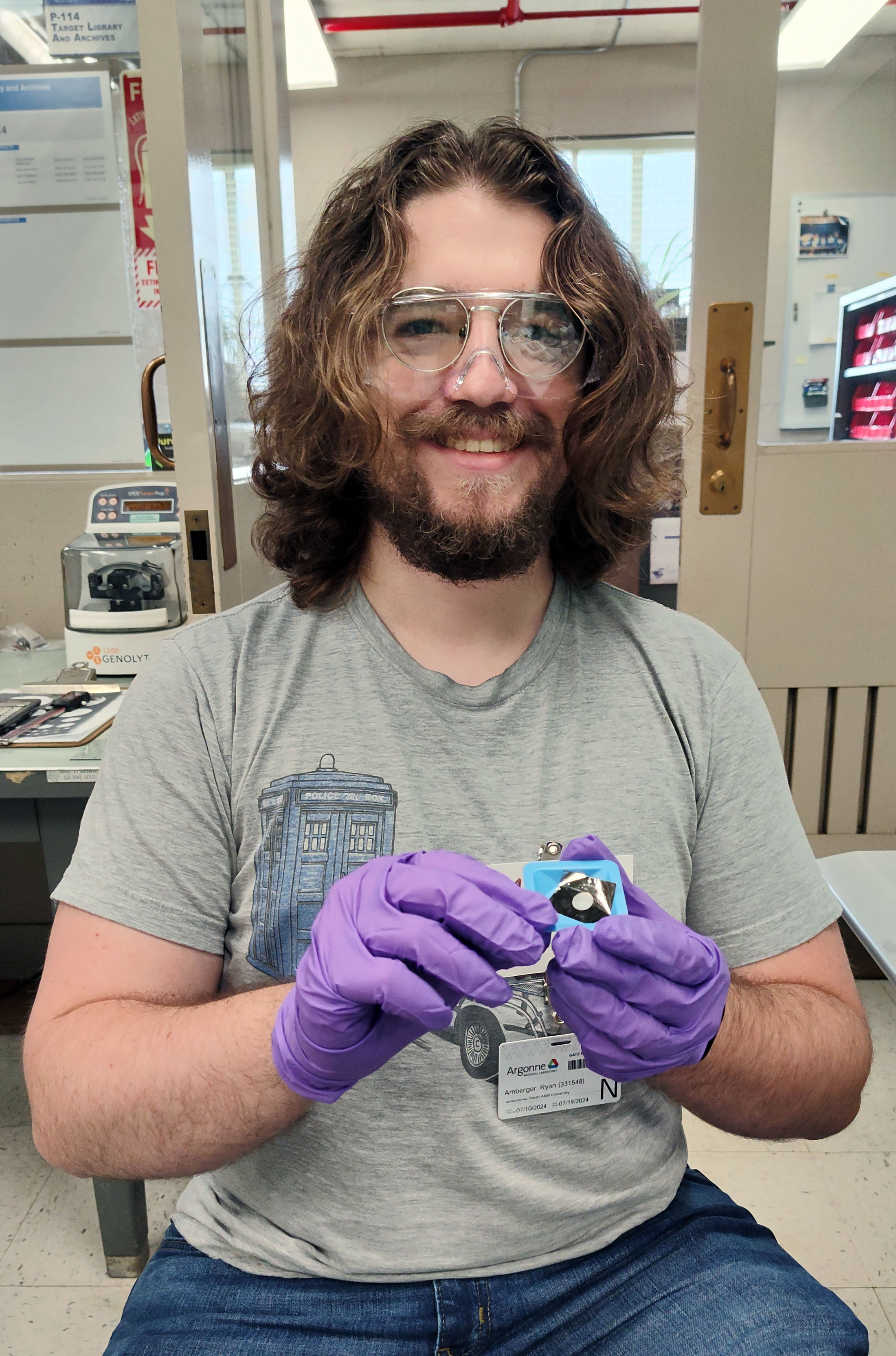 Texas A&amp;M University physics graduate student Ryan Amberger smiles while sitting in a laboratory at Argonne National Laboratory, where he is wearing goggles and purple latex gloves while holding a component of a target involved in nuclear physics experiments.