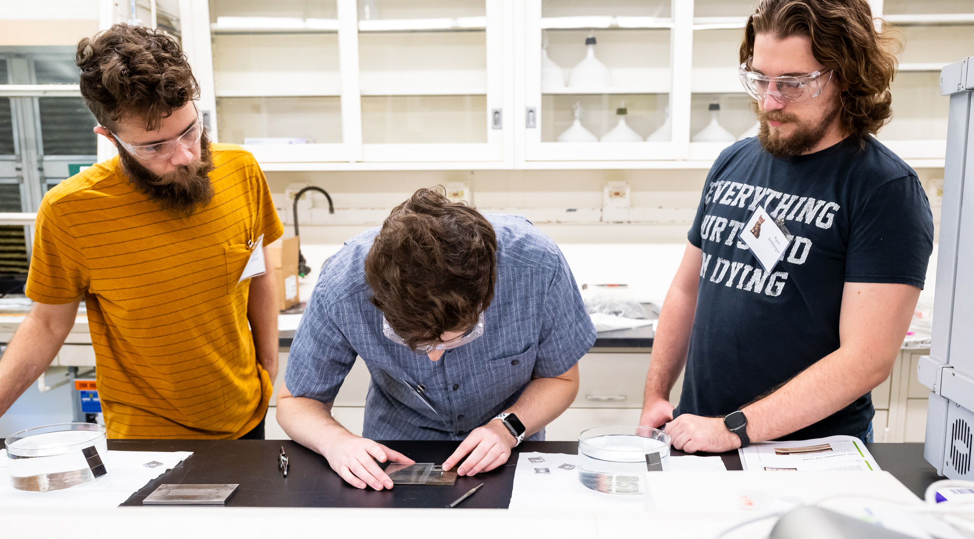 Three people including Texas A&amp;amp;M University physics graduate student Ryan Amberger at right wear goggles while standing at a lab table, where the middle person aligns square-shaped target samples as the other two observe.