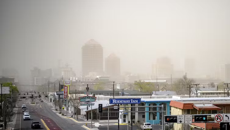 A hazy view of Albuquerque, showing a nearly empty street with traffic lights, several small businesses including a Rodeway Inn, and the distant outline of downtown buildings obscured by fog.