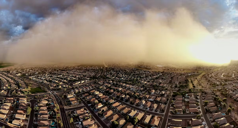 A large dust storm approaching a suburban area with numerous identical houses, under a cloudy sky.