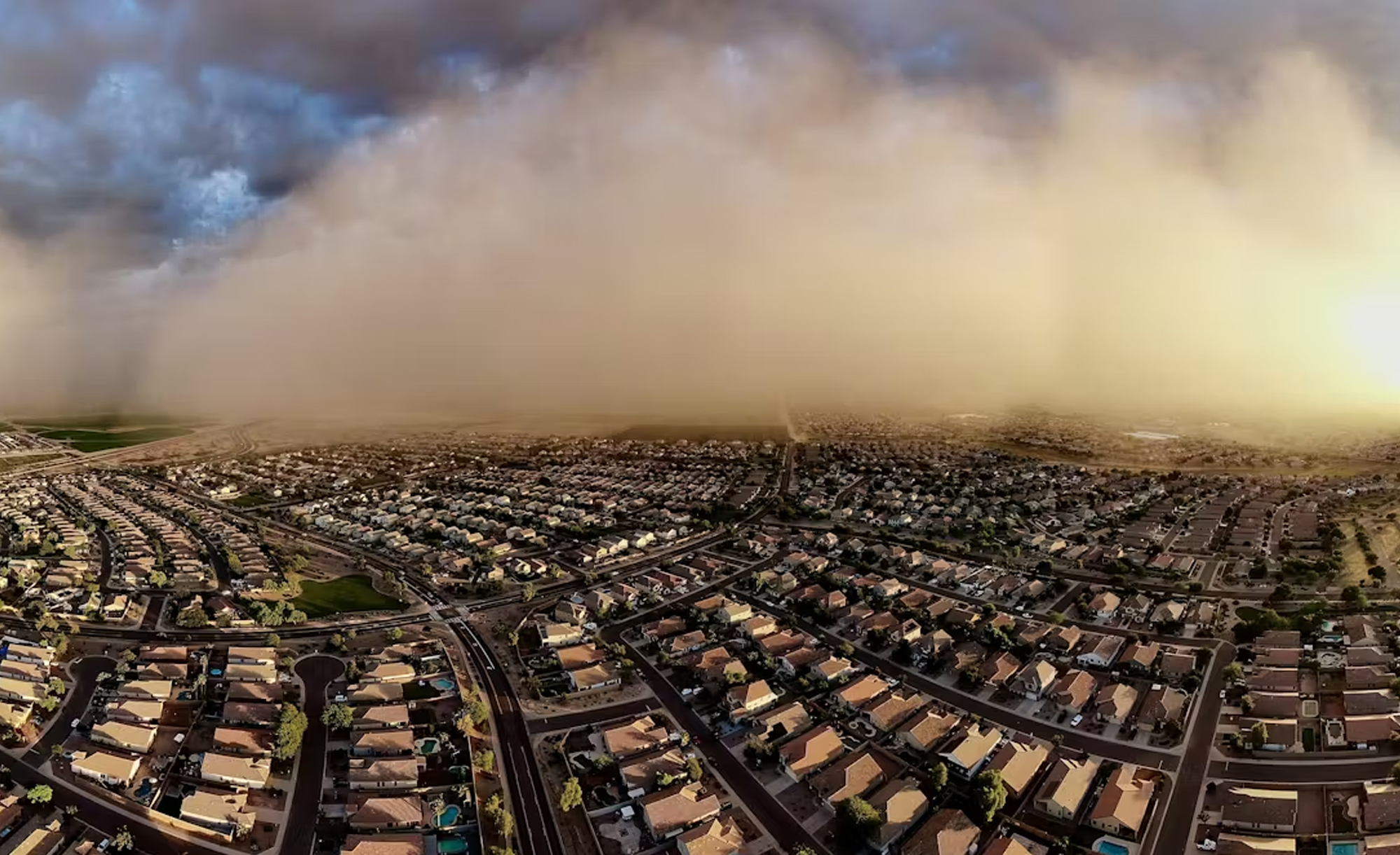 A large dust storm approaching a suburban area with numerous identical houses, under a cloudy sky.