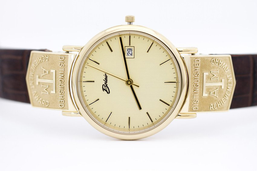 A Boucheron wristwatch with a gold case and a beige dial featuring a date window, set against a white background. The watch is attached to a brown leather strap with embossed text reading "Distinguished Achievement Award.