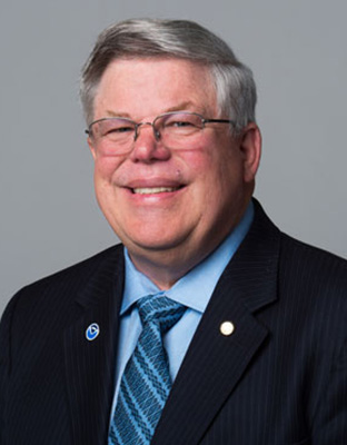 Headshot of Richard Feely wearing a black suit and blue shirt and tie, against a grey backdrop.