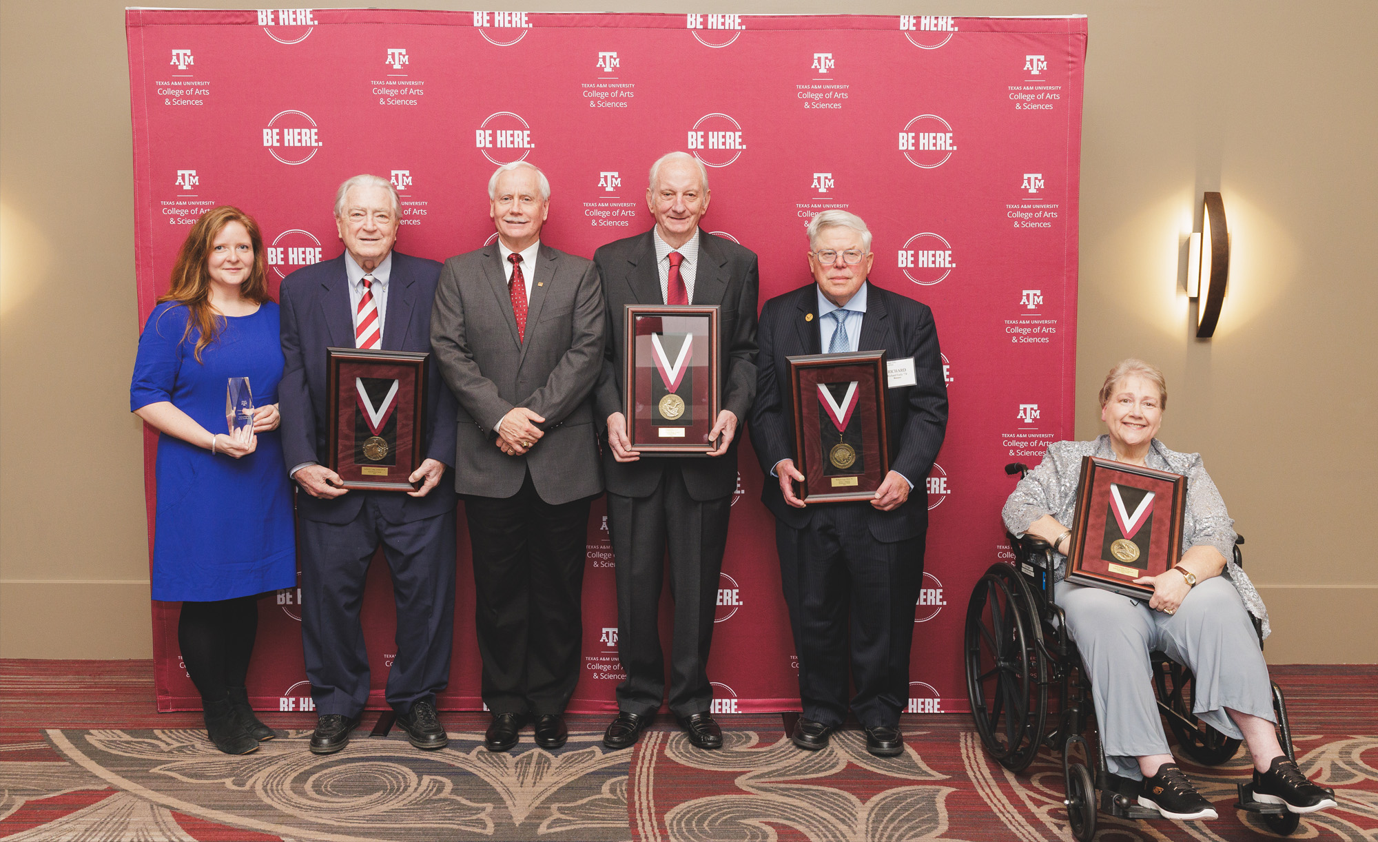 Group of six individuals holding awards, standing in front of a Texas A&amp;M University banner with the slogan "Be Here.