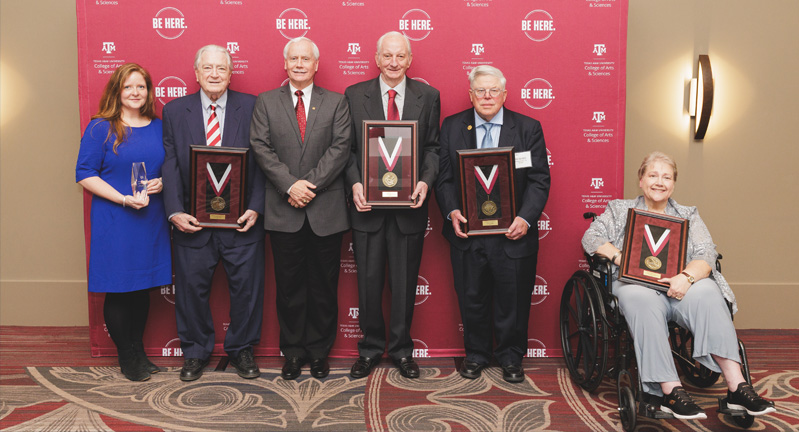 Group of six individuals holding awards, standing in front of a Texas A&amp;M University banner with the slogan "Be Here.