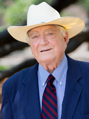 Luke Soules, wearing a blue suit and cowboy hat, stands in front of a natural backdrop.