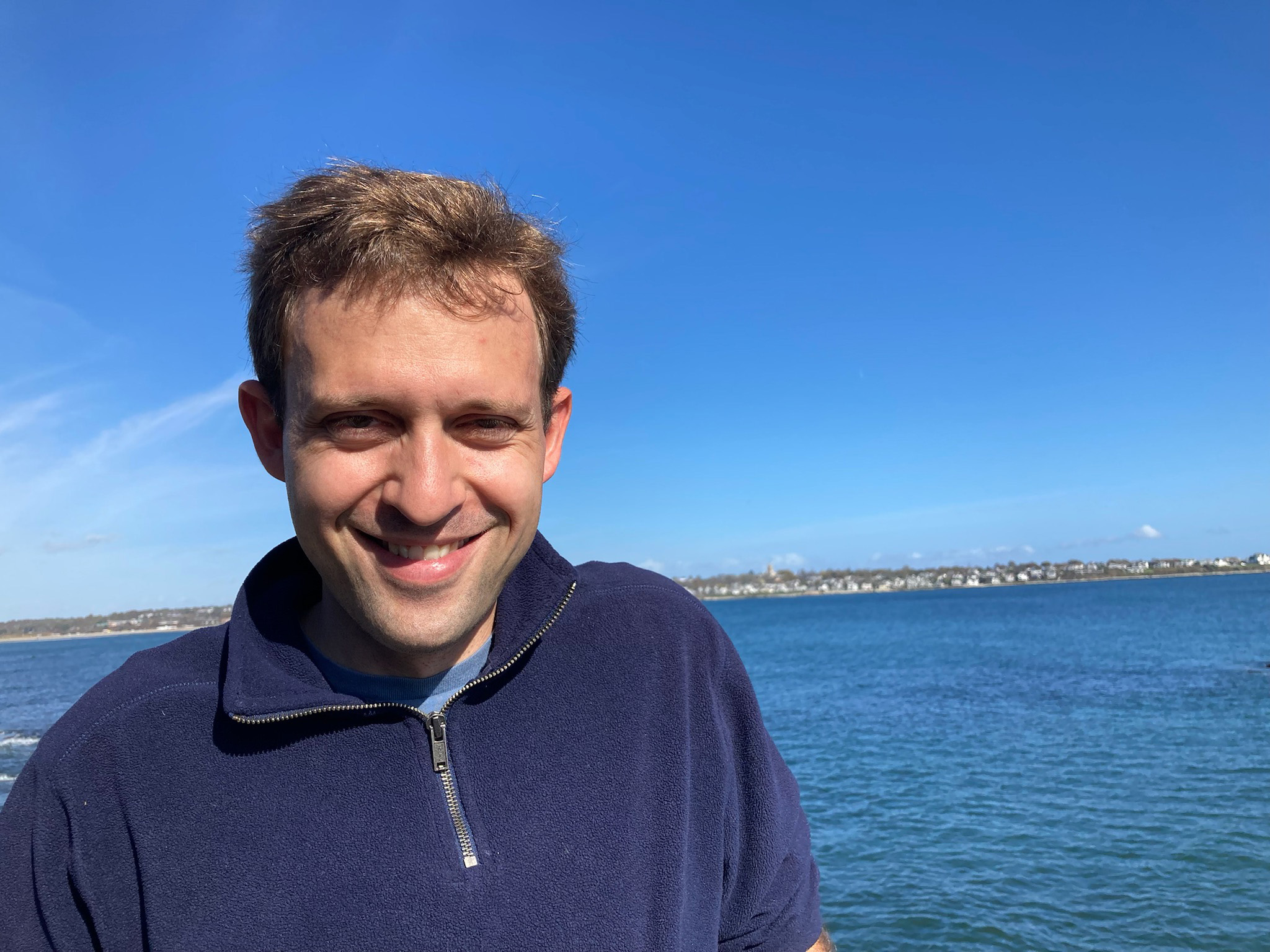 Smiling person in a navy blue top standing in front of a body of water with a clear blue sky and a distant city skyline.