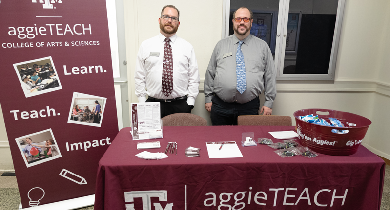 Kenric Davies and Colby Richardson standing behind a promotional table for the AggieTEACH program at Texas A&amp;M University, flanked by banners displaying the university's logo and the College of Arts &amp; Sciences. The table features brochures and promotional items.