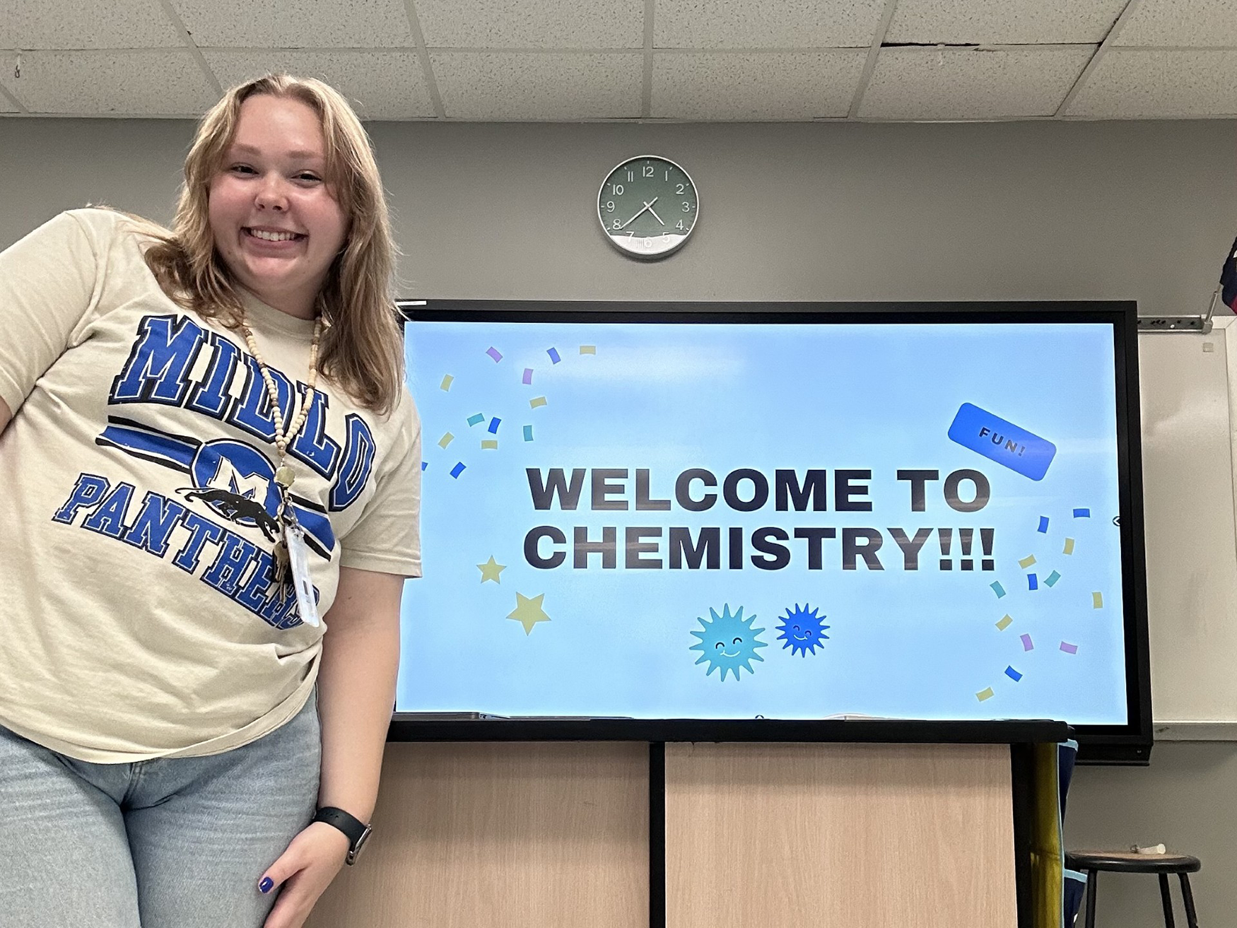 Denae Jaksik in a Midland Panthers t-shirt smiling beside a screen displaying a 'Welcome to Chemistry!!!' message with festive confetti graphics.