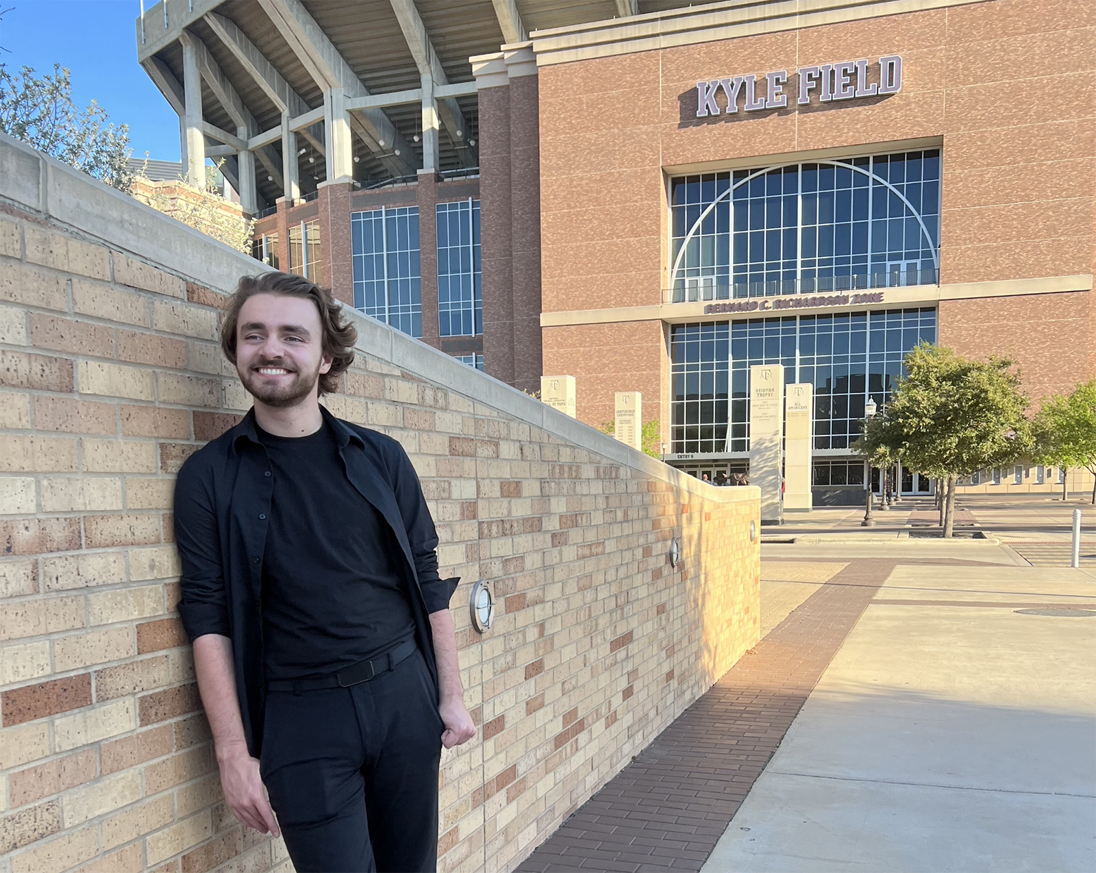Matthew Morris in a black outfit smiling and leaning against a brick wall outside Kyle Field stadium.
