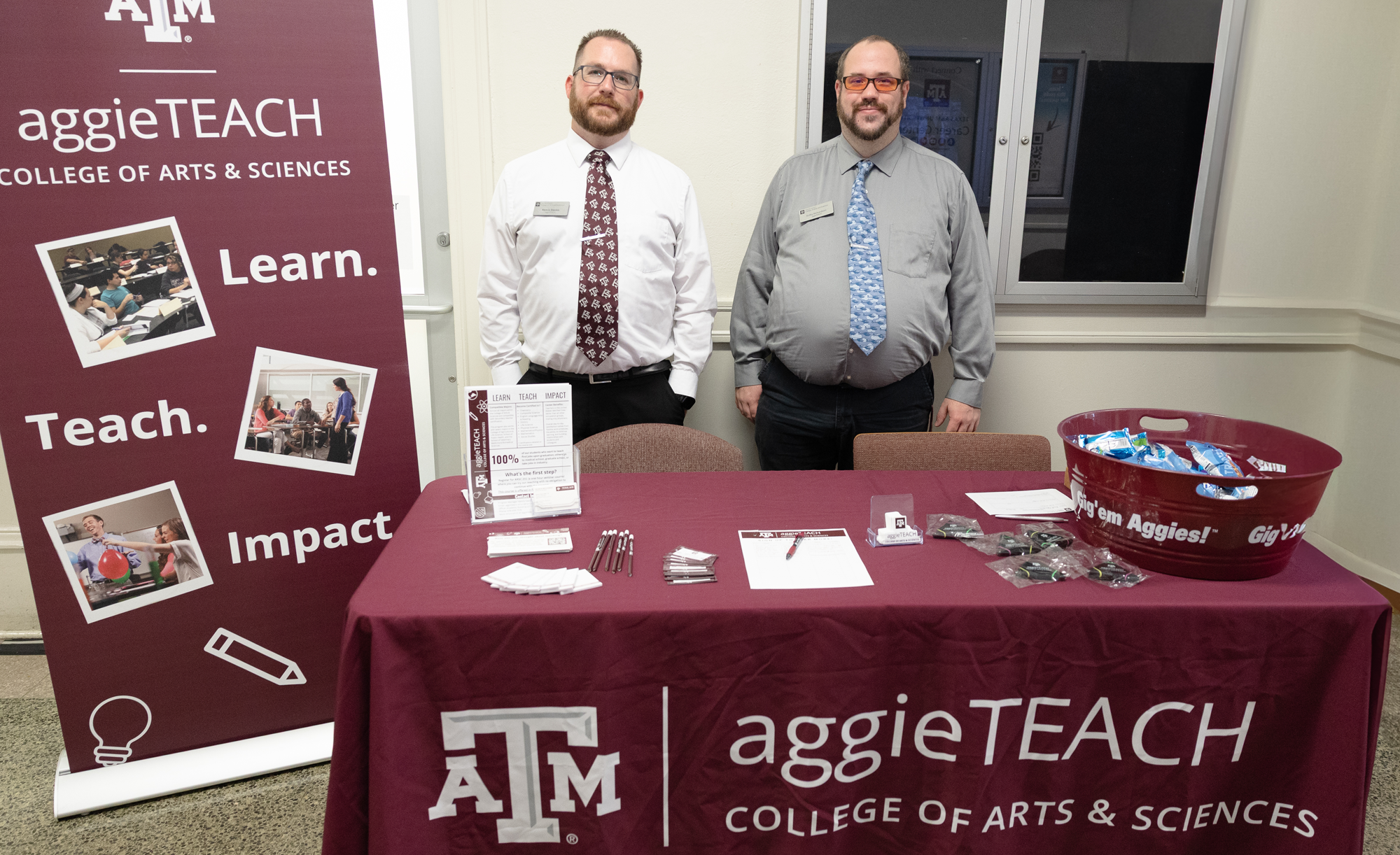 Kenric Davies and Colby Richardson standing behind a promotional table for the AggieTEACH program at Texas A&M University, flanked by banners displaying the university's logo and the College of Arts & Sciences. The table features brochures and promotional items.