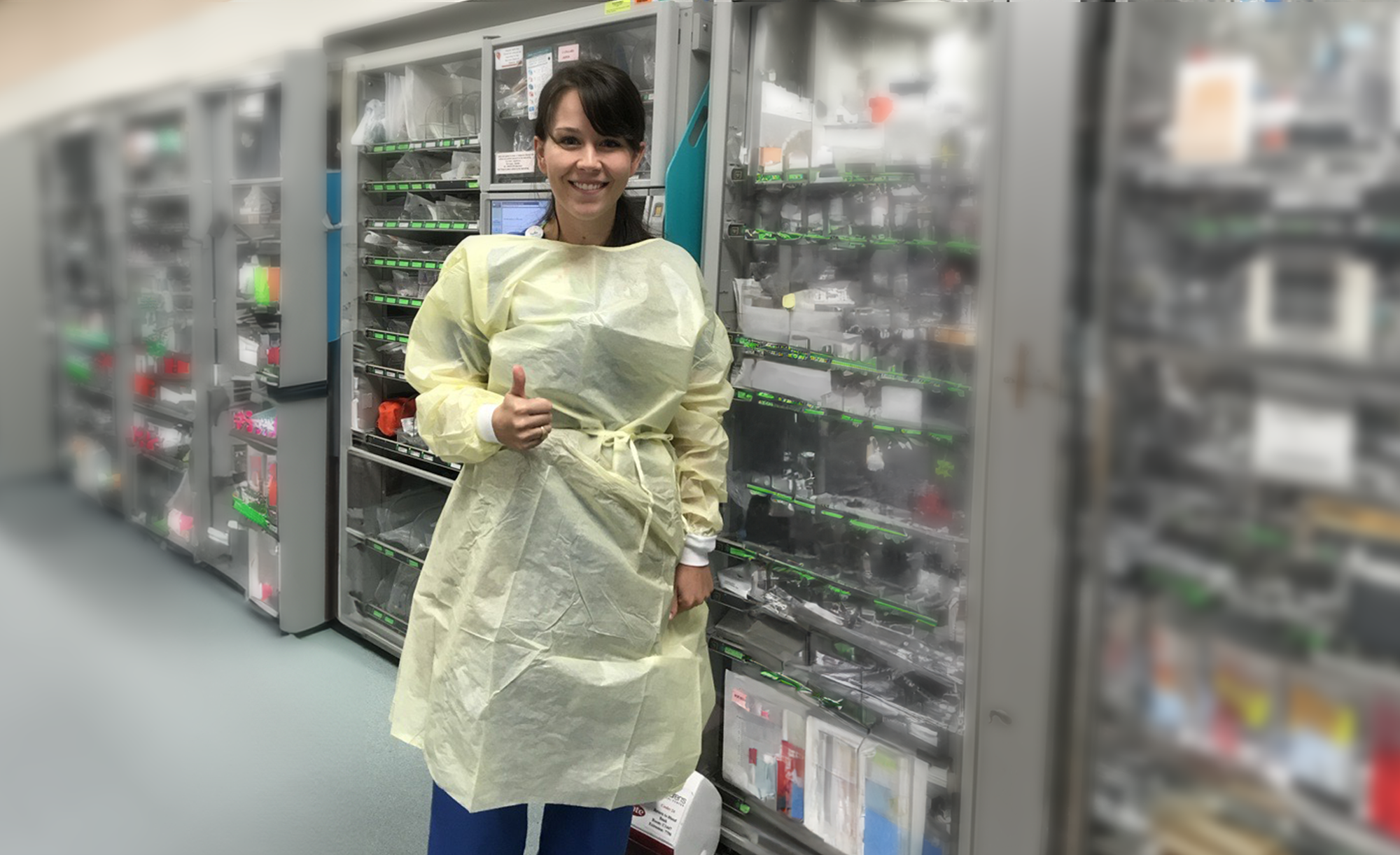 Kaitlin Tindel in a lab coat smiling and giving a thumbs up in front of hospital racks.