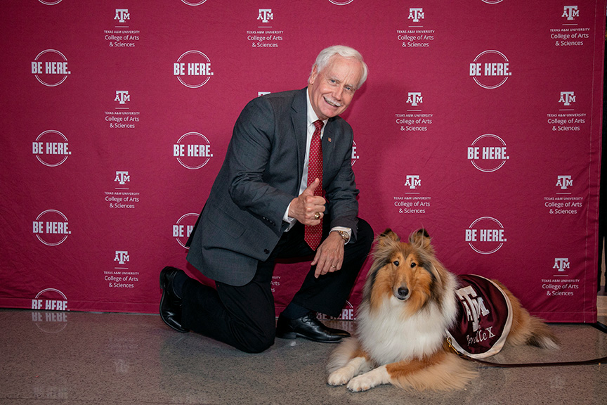 Person kneeling next to Reveille, the Texas A&M University mascot, in front of a backdrop with the university's logos and the phrase "Be Here." Both are looking at the camera.