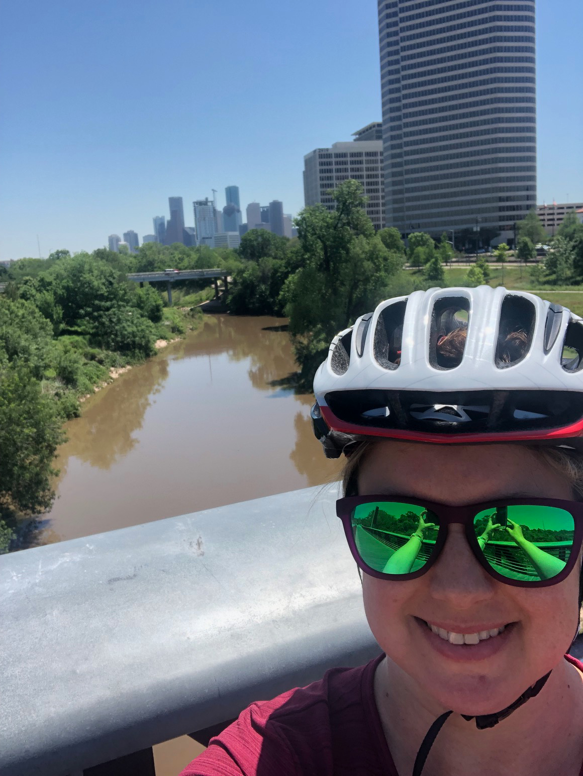 Selfie of Aubin Phillips wearing a bike helmet and sunglasses with Houston skyline and a river in the background.