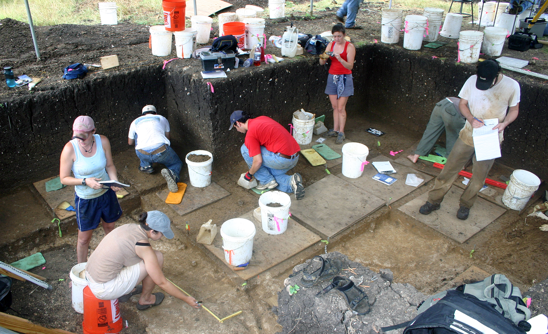 Archaeological dig in progress with several individuals examining and documenting different sections of an excavation site.