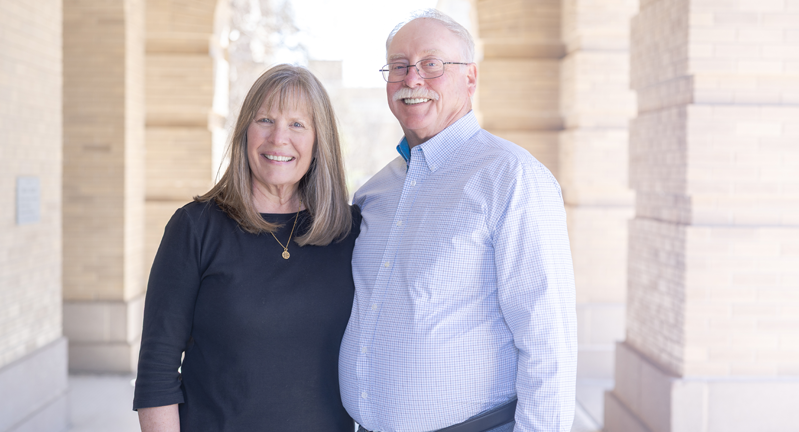Dennis (right) and Melinda Hughes smiling and standing close together in front of a building with columns.
