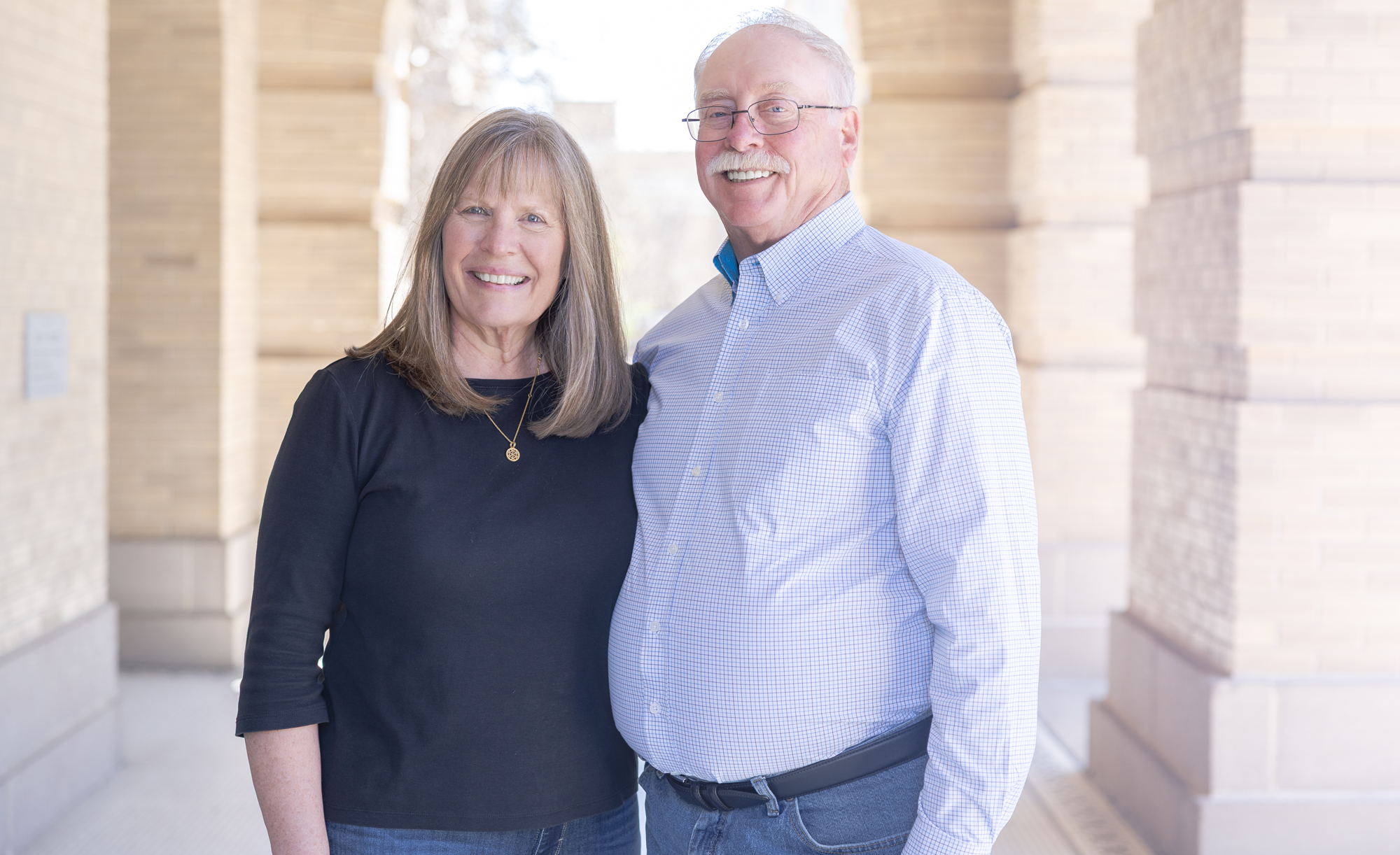 Dennis (right) and Melinda Hughes smiling and standing close together in front of a building with columns.