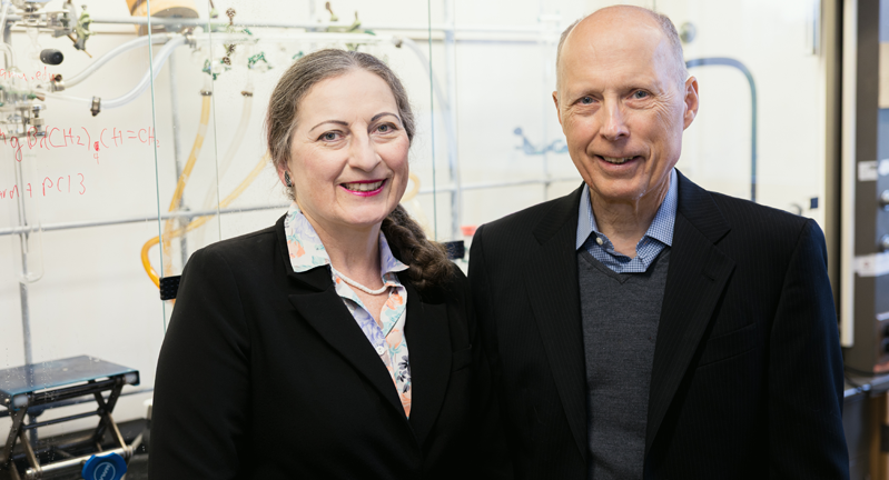 Dr. Janet Bluemel (left) and Dr. John Gladysz  smiling in a laboratory with chemical formulas written on the glassware in the background.