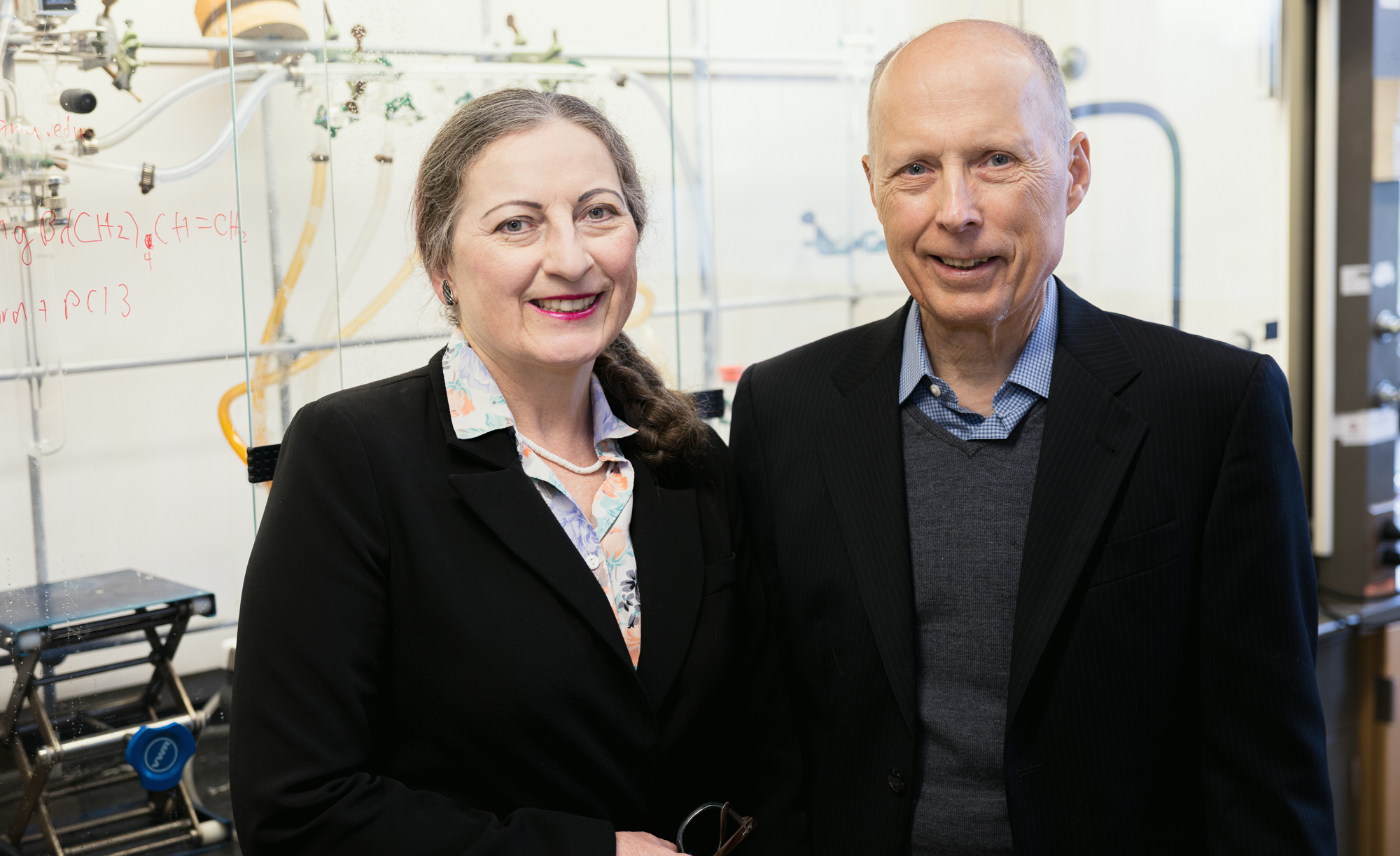 Dr. Janet Bluemel (left) and Dr. John Gladysz smiling in a laboratory with chemical formulas written on the glassware in the background.