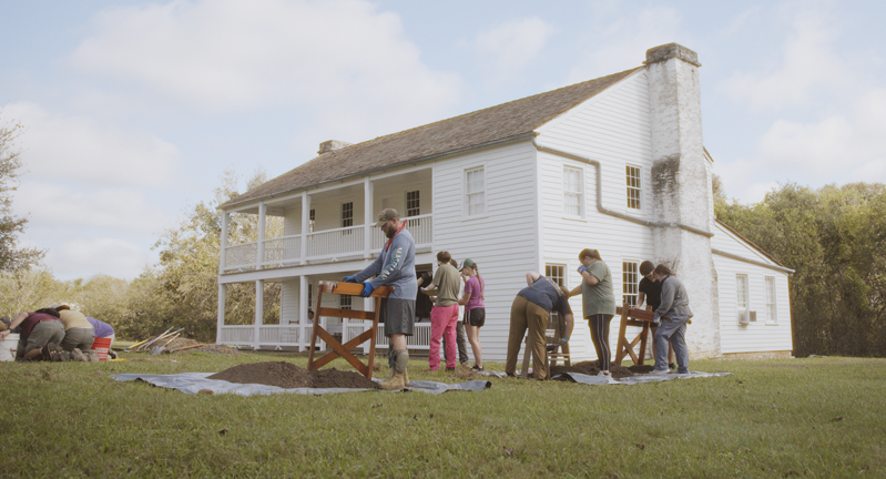 A group of people of various ages participating in an outdoor gardening activity near a large white house with a chimney.