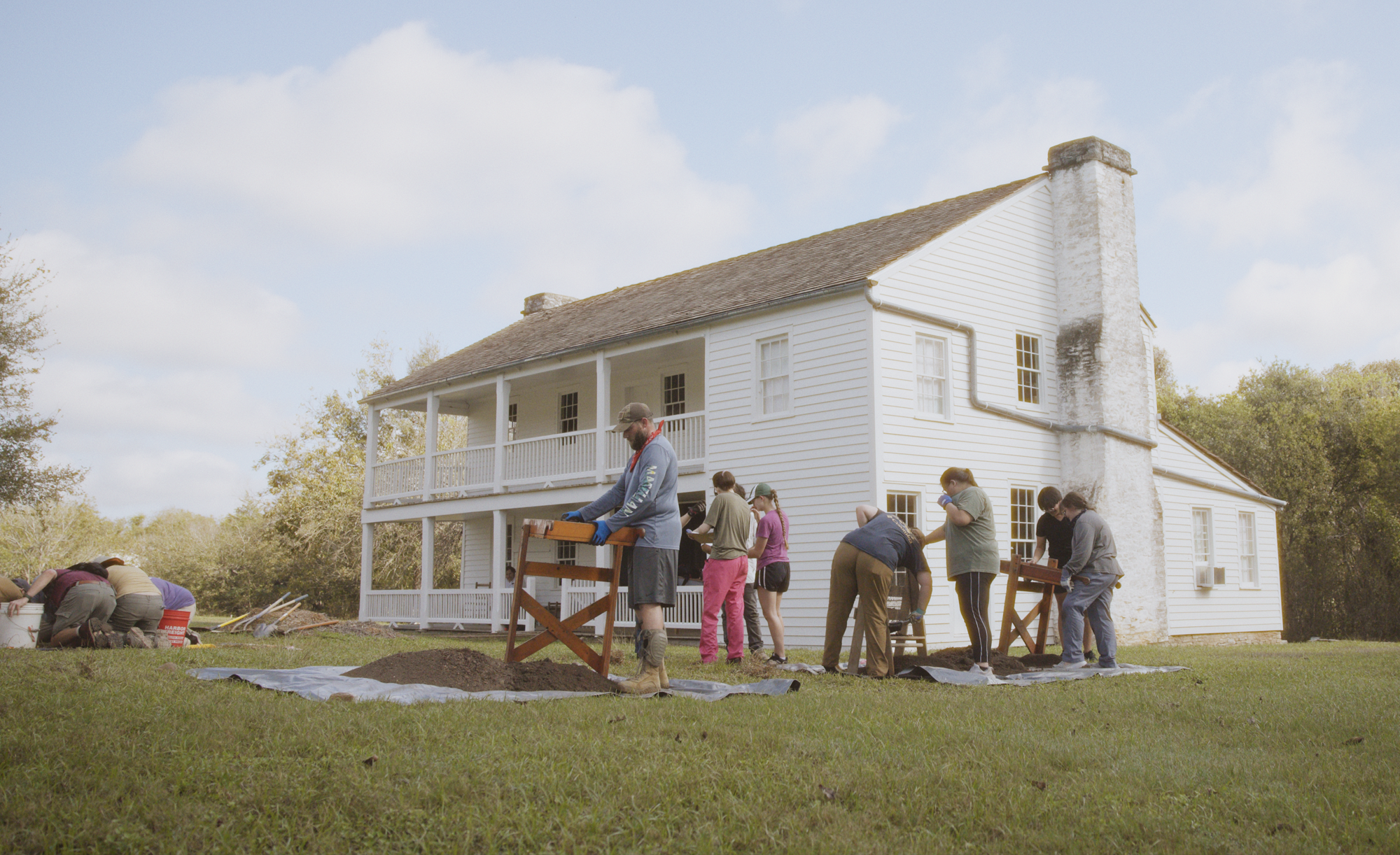 A group of people of various ages participating in an outdoor gardening activity near a large white house with a chimney.
