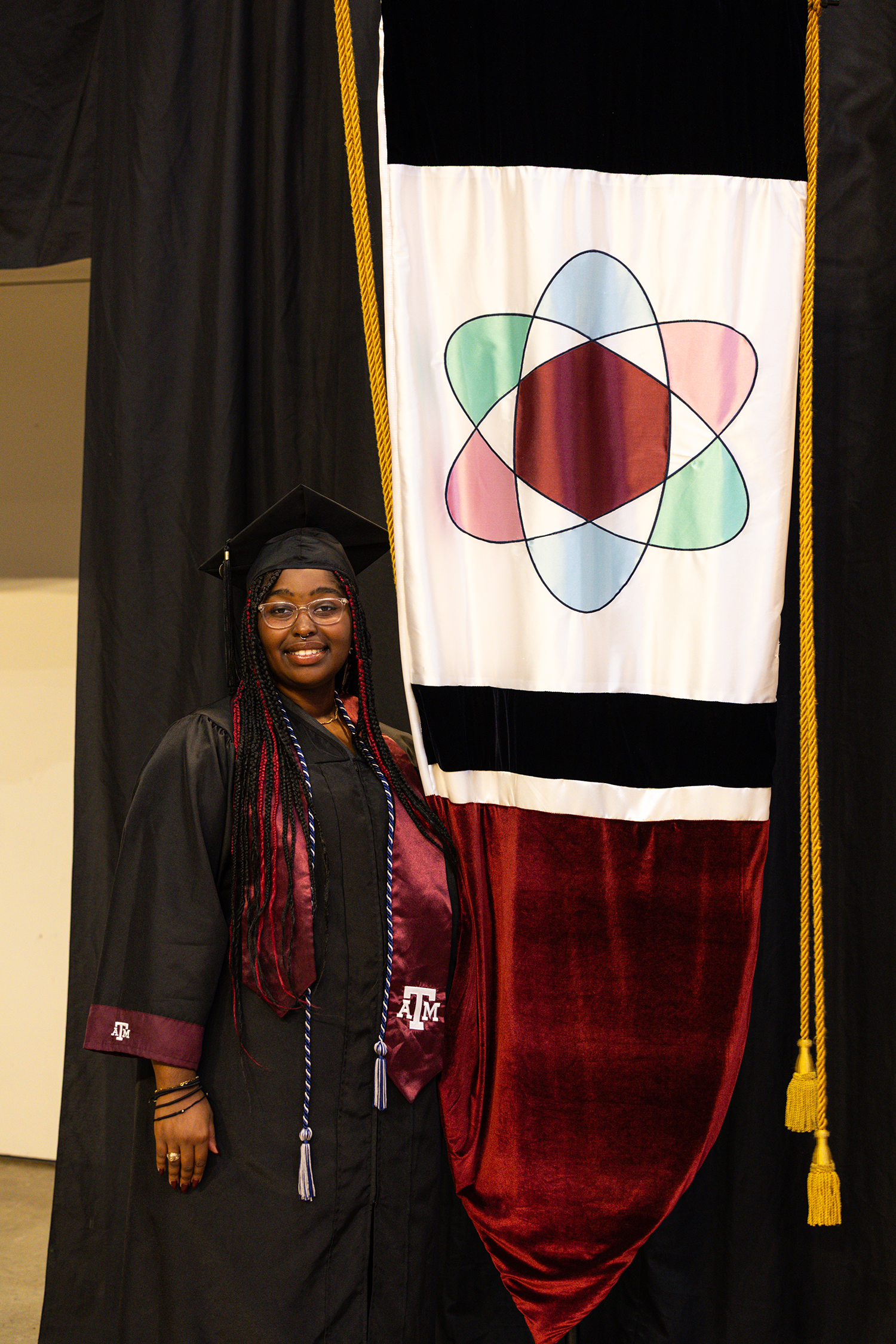 A graduate in cap and gown stands beside a Texas A&M University banner, smiling proudly.