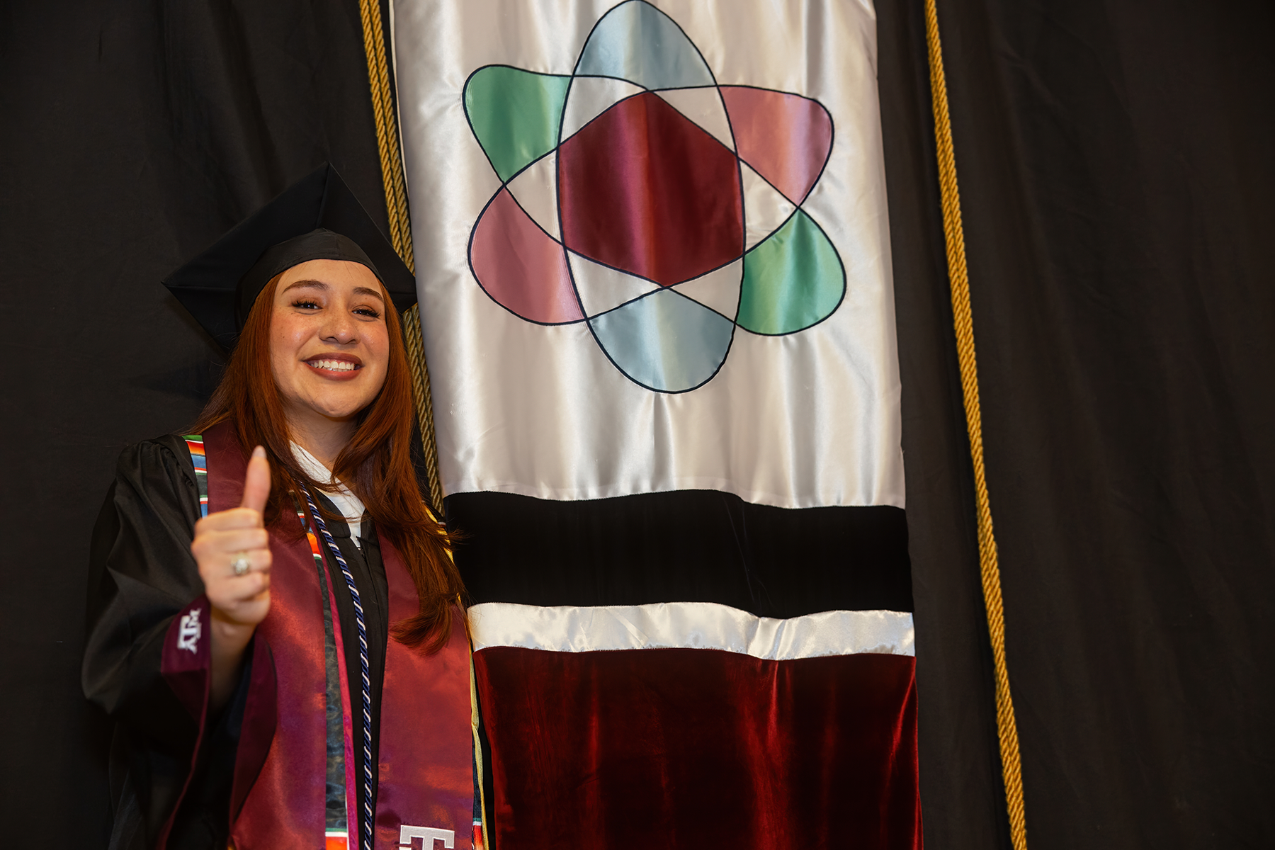 Graduate in cap and gown giving a thumbs up next to a flag with a multicolored atom symbol, representing Texas Tech University.