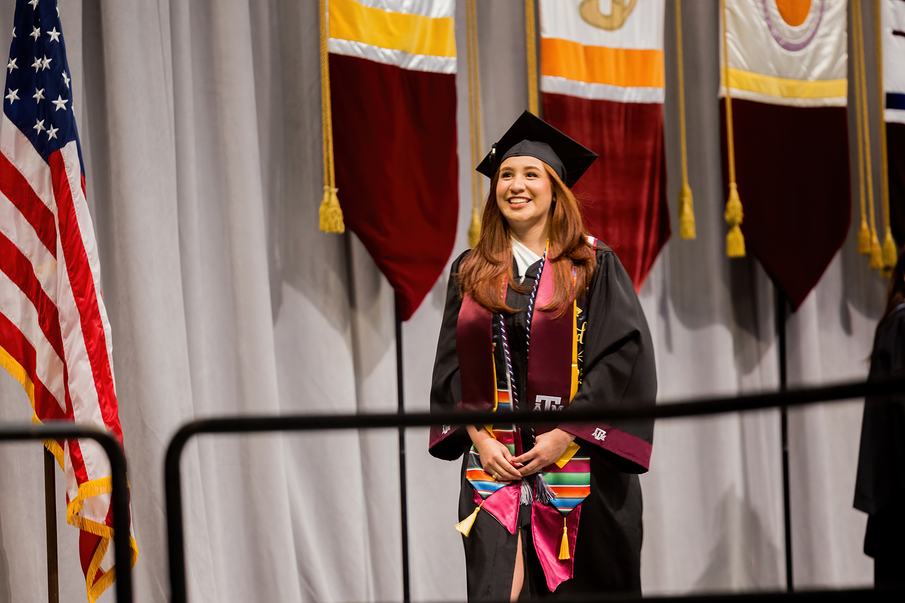 A graduate smiling at a university ceremony, holding a diploma and wearing a cap, gown, and various honor cords. U.S. and state flags, along with other banners, are displayed in the background.