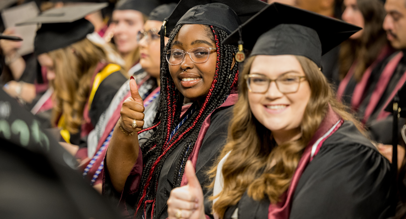 Graduates in caps and gowns giving a thumbs up at a graduation ceremony.