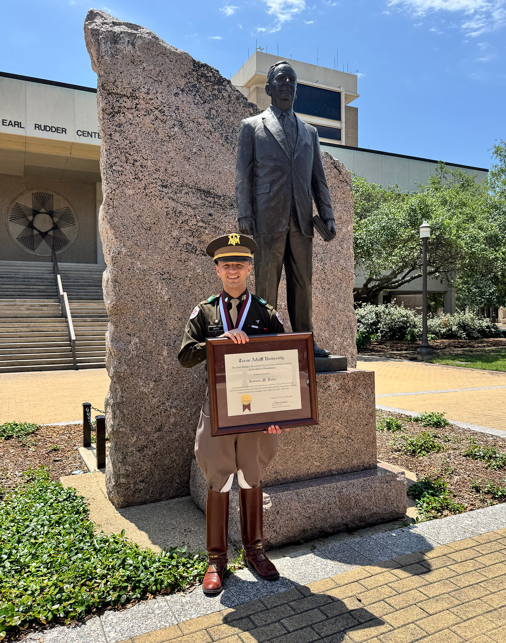 Jackson Baker in military uniform holding a framed certificate stands in front of the James Earl Rudder statue at Texas A&M University.