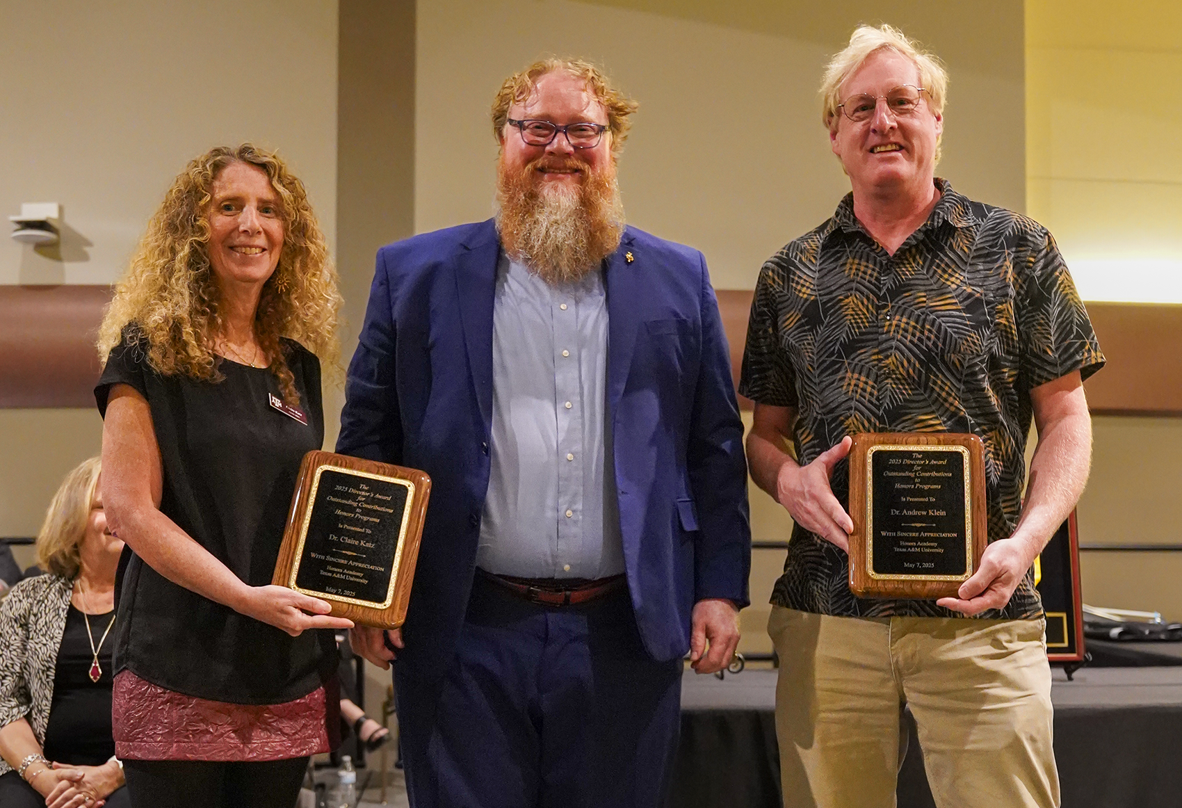 Three adults standing on stage, each holding an award plaque. The person in the center wears a blue suit, flanked by two individuals, one in a patterned blouse and black pants, and the other in a tropical print shirt. They are smiling and appear at a formal award ceremony.