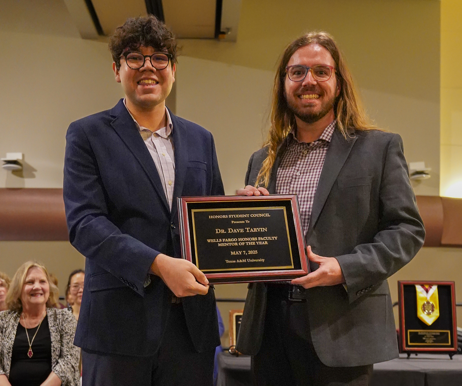 Two individuals smiling and holding an award plaque at an indoor ceremony. One is dressed in a navy suit and the other in a checked shirt and grey blazer. The plaque reads "Dr. Davis Taylor Award for Teaching Excellence.