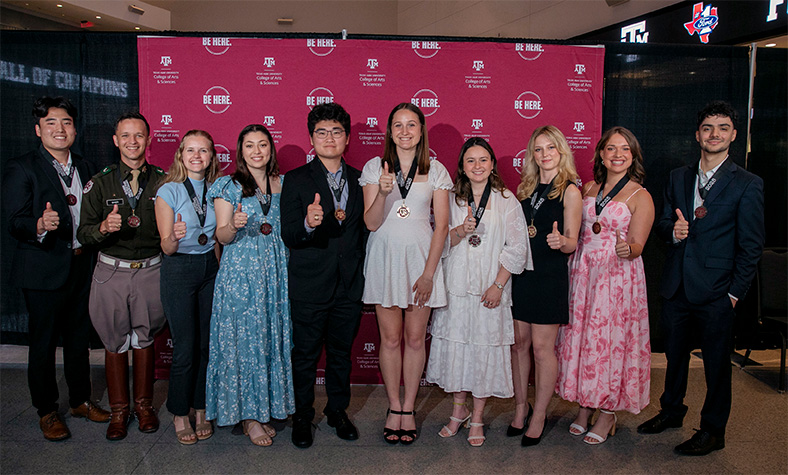 Group of ten Texas A&M University students displaying thumbs-up at an event, standing in front of banners with university logos.