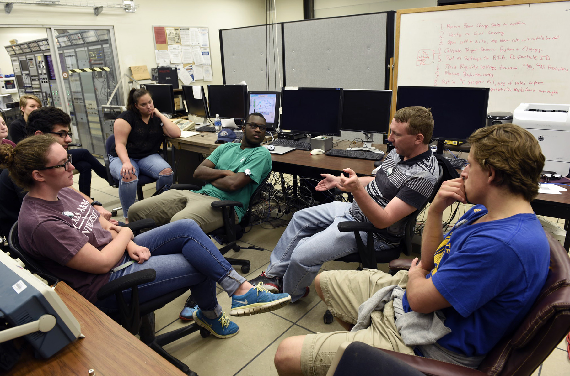 A group of undergraduate students sit in chairs beside rows of tables while listening to Cyclotron Institute accelerator physicist Brian Roeder during a group meeting.
