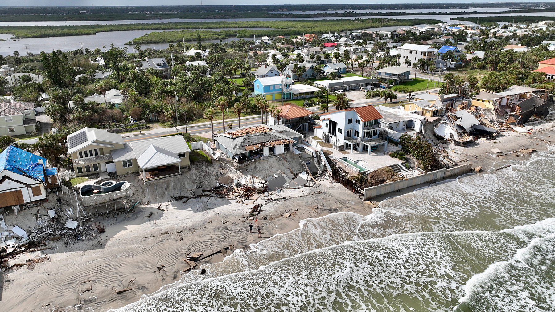 Aerial view of coastal damage with several homes and buildings destroyed along the shoreline, reflecting the impact of a severe storm. Waves continue to lap against the compromised coastline.
