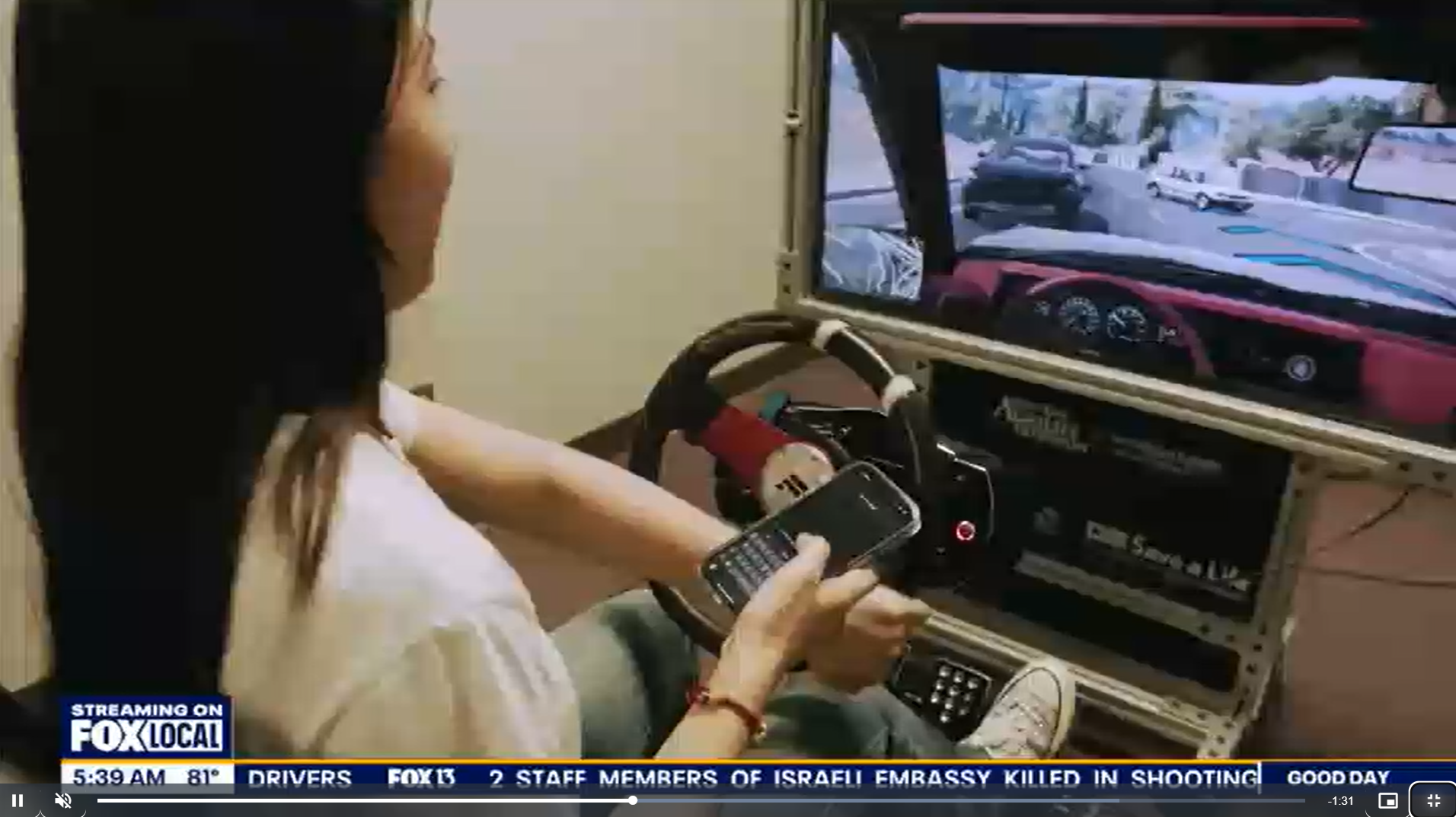 Person using a driving simulator with a view of a road on the monitor, FOX Local news banner visible at the bottom.