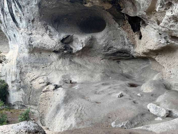 View of a large, naturally eroded cave with sandy floors surrounded by rocky walls, with sparse vegetation visible at the edges.