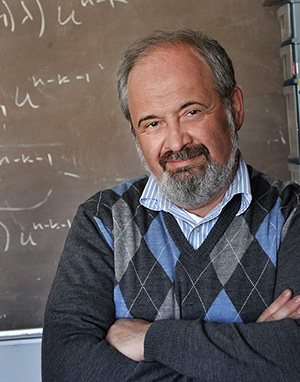 Portrait of Dr. Peter Kuchment, standing in front of a chalkboard filled with mathematical formulas, smiling at the camera. Kuchment is wearing a blue and gray argyle sweater vest over a checked shirt.