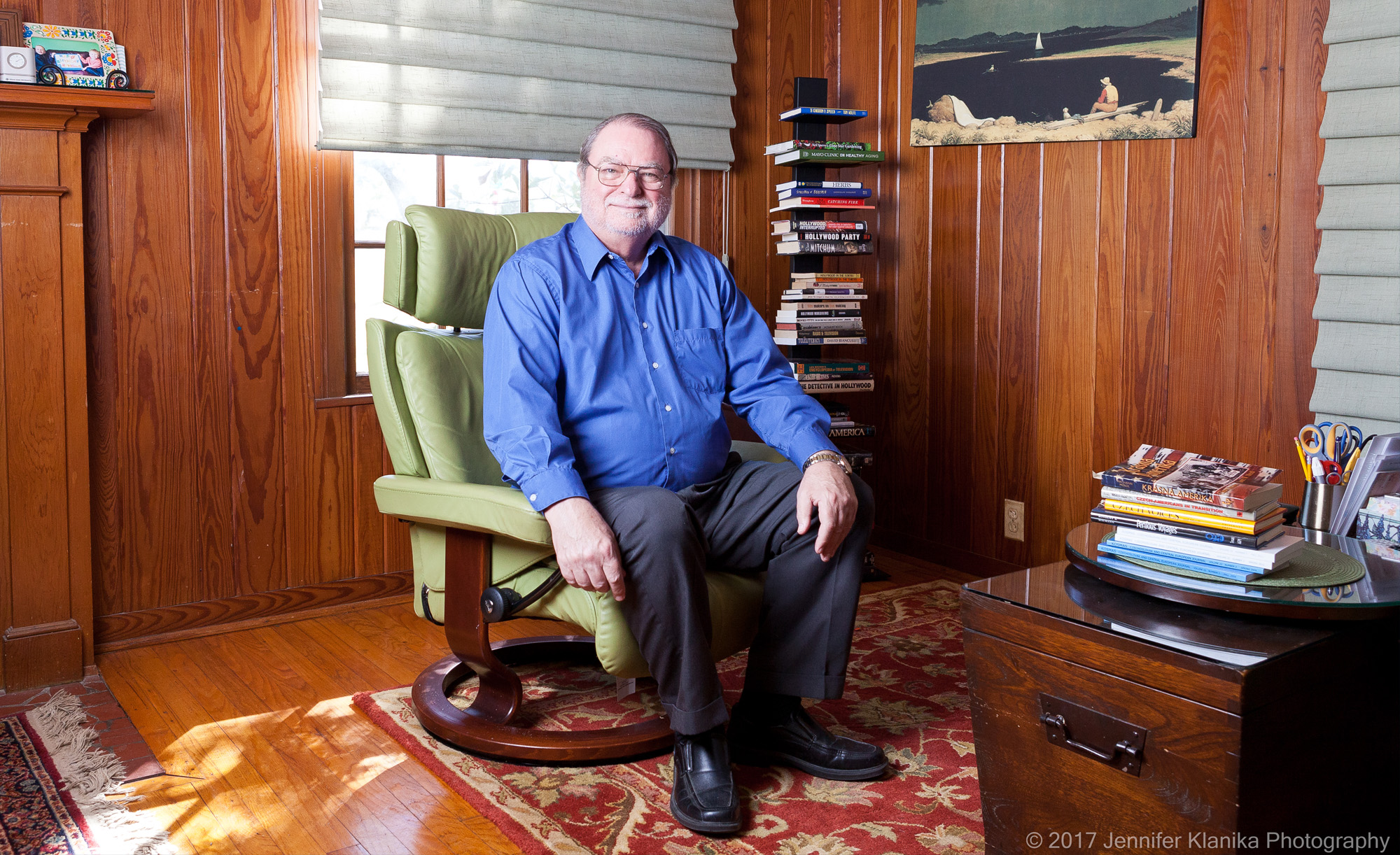 An individual seated in a green armchair in a wood-paneled room, with a stack of books to one side and a painting above. A toy car is on a shelf in the background.